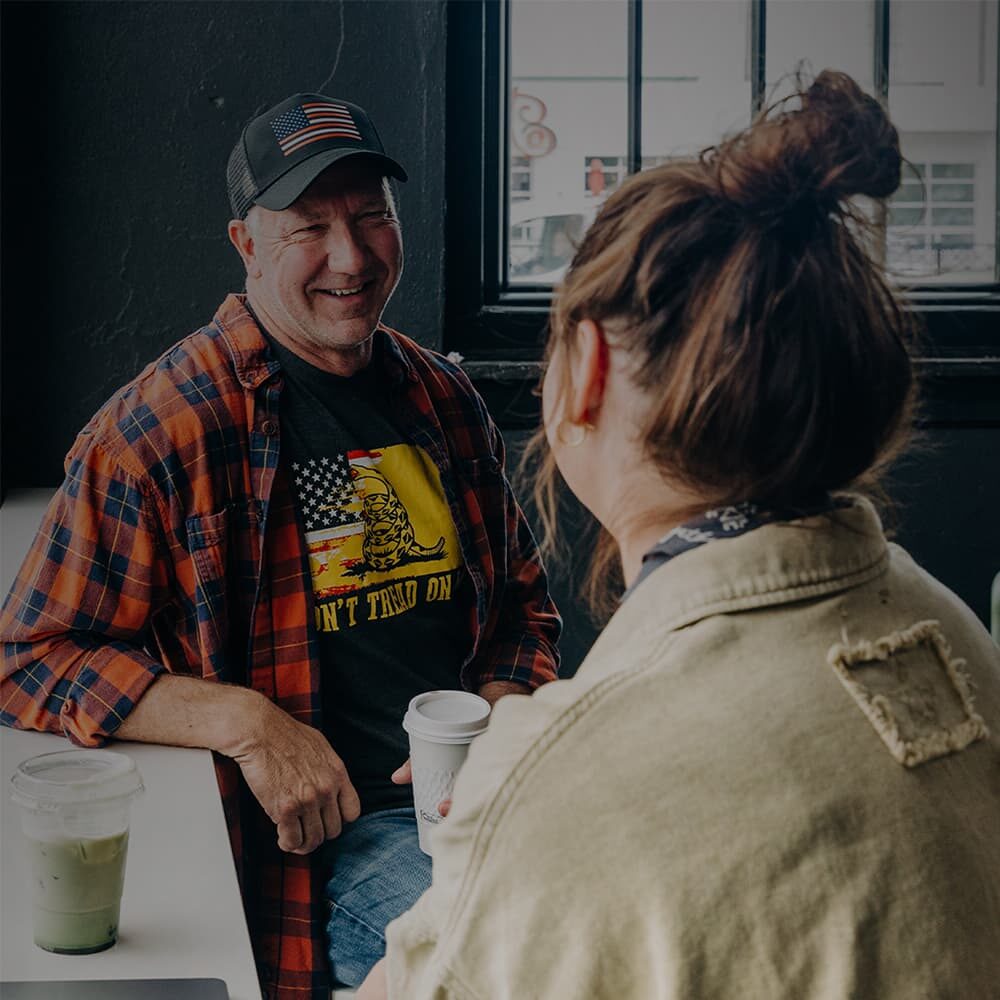 An older man wearing an American flag t-shirt and a flannel shirt is having a friendly conversation with a woman wearing a bun hair style.