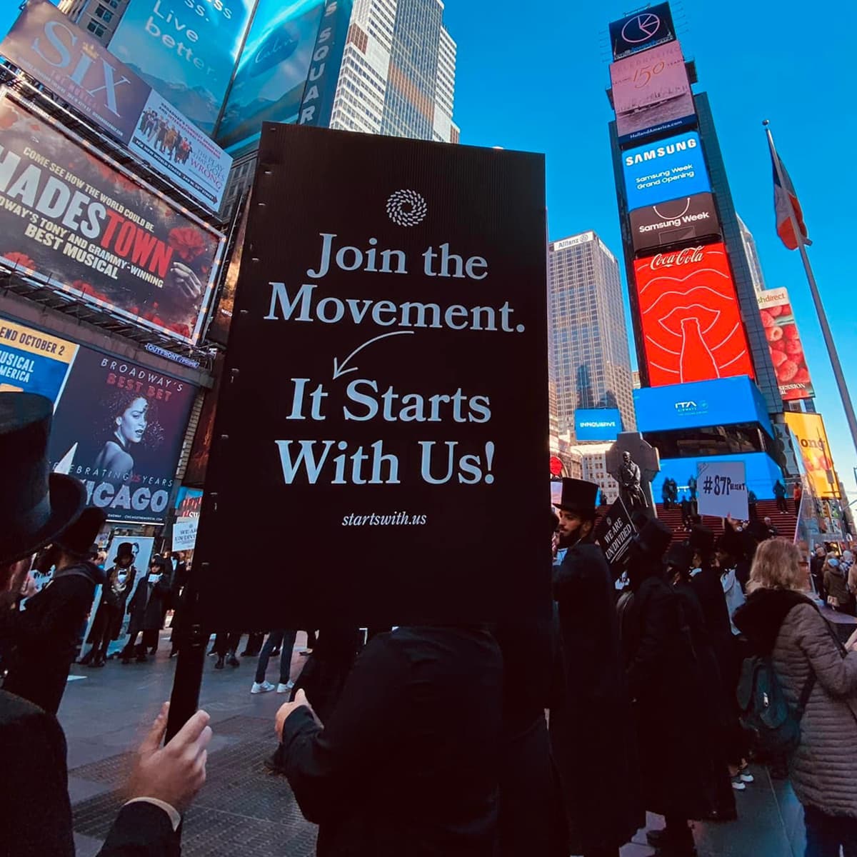 A group of people dressed as Abraham Lincoln in Times Square holding a sign that says, "Join The Movement. It Starts With Us!"